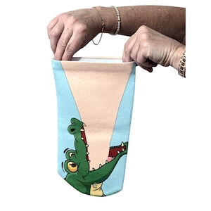 Puede incluir: Una bolsa de tela rectangular con un fondo azul claro y una ilustración gráfica de un cocodrilo verde con la boca abierta. El cocodrilo tiene ojos amarillos grandes y dientes blancos, situado en la parte inferior de la superficie de tela azul.