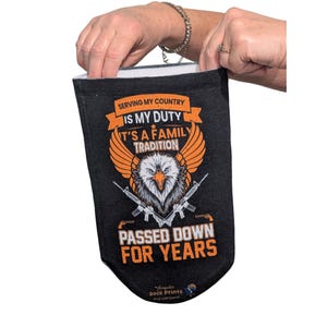 Puede incluir: Calcetín negro con el texto "SERVING MY COUNTRY IS MY DUTY IT'S A FAMILY TRADITION PASSED DOWN FOR YEARS" y un águila con rifles cruzados. El calcetín está abierto por una persona.