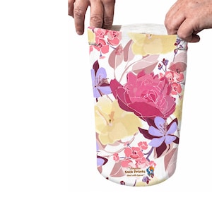 May include: A cylindrical fabric prosthetic sock featuring a white background with a vibrant floral pattern in shades of pink, yellow, and light purple. The lower portion displays the text Amputee Sock Prints Heal with Appeal.