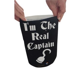 May include: A black cylindrical beverage insulator sleeve featuring the white text "I'm The Real Captain" printed in a bold, stylized font. Below the text is a white graphic illustration of a pirate hook. The sleeve has a white interior lining.