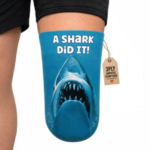 May include: A blue amputee stump sock featuring a graphic of a shark with its mouth open and the text A SHARK DID IT! in white letters. A brown cardboard tag attached with a string reads 3PLY AMPUTEE STUMP SOCK with a small red heart icon.