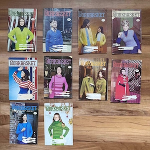 May include: A collection of vintage "Workbasket" magazines, each featuring colorful knitwear on the cover. The covers showcase various styles of sweaters, cardigans, and other knitted garments, with models posing in front of different backgrounds. The magazines are displayed on a wooden surface.