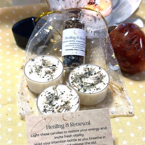May include: A collection of spiritual items, including three small candles topped with herbs, a small glass bottle with a label that reads "Healing & Renewal", and a card with the same text. The items are arranged on a yellow surface.