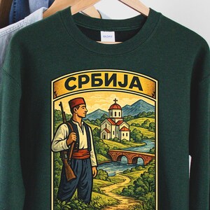 Puede incluir: Una sudadera de manga larga de color verde oscuro con un estampado gráfico de una persona con vestimenta tradicional que sostiene un rifle, cerca de una iglesia, un puente de piedra y un río. El texto "СРБИЈА" aparece en letras cirílicas.