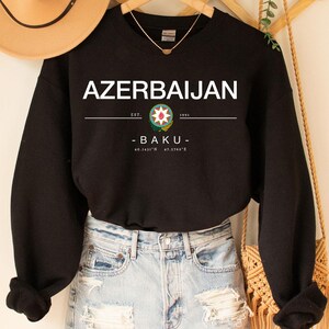 Könnte beinhalten: Schwarzes Sweatshirt mit dem Wort "AZERBAIJAN" in weißen Blockbuchstaben. Darunter eine Grafik und das Wort "BAKU". Das Sweatshirt wird mit Jeansshorts und einem beigen Hut kombiniert.