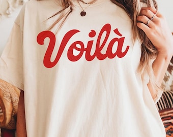 Op de afbeelding: Crèmekleurig T-shirt met het woord "Voilà" in rode letters. Het shirt heeft een losse pasvorm en is gemaakt van een zacht materiaal. De tekst staat in het midden van de voorkant van het shirt.