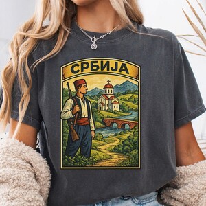 Puede incluir: Una camiseta color pimienta con una ilustración gráfica de una persona con vestimenta tradicional sosteniendo un rifle, frente a un paisaje con una iglesia y un puente, con el texto cirílico "СРБИЈА" impreso en un arco amarillo arriba.