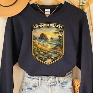 Peut inclure: Un sweat-shirt bleu marine avec un motif de Cannon Beach, Oregon. Le motif est une scène de paysage détaillée avec le texte "CANNON BEACH" et "OREGON". Le sweat-shirt est associé à un short en jean et un chapeau beige.