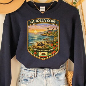 May include: Navy blue sweatshirt with a La Jolla Cove graphic. The graphic features a coastal scene with a sunset, ocean, and palm trees. The sweatshirt is paired with denim shorts and a tan hat.