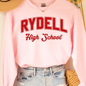 Può includere: Felpa rosa con la scritta "Rydell High School" in lettere rosse. La felpa è appesa a un supporto in legno con un cappello di paglia e una borsa intrecciata.