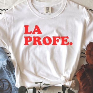 May include: White t-shirt with the text "LA PROFE." in red letters. The shirt is laid out on a striped surface with a pair of brown boots and blue jeans.
