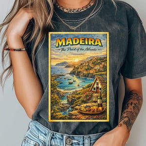 May include: Dark gray t-shirt featuring a colorful travel poster for Madeira, Portugal. The poster depicts a coastal landscape with buildings, boats, and the text "MADEIRA - The Pearl of the Atlantic". The shirt is made of a soft material.
