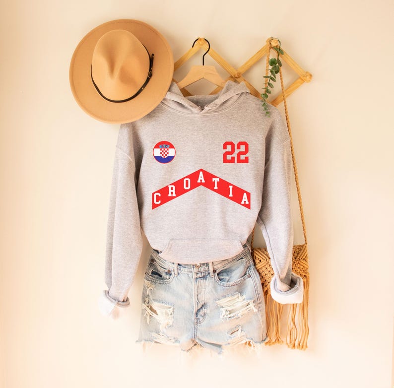 May include: Gray hooded sweatshirt with a red and white Croatian flag, the word "CROATIA" in red letters, and the number "22" in red. The sweatshirt is hanging on a wooden rack with a straw bag.