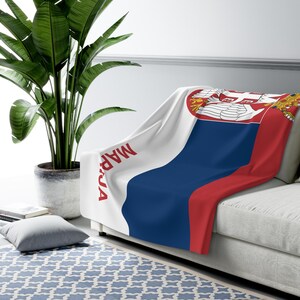 Puede incluir: Una manta con la bandera serbia en blanco, azul y rojo, colocada sobre un sofá. La manta presenta el escudo de armas serbio y la palabra "MAPJA" en rojo. Una gran planta verde en una maceta gris se encuentra en el fondo.