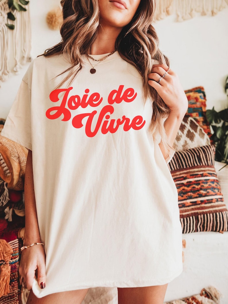 May include: A cream-coloured t-shirt featuring the phrase "Joie de Vivre" in red, cursive lettering. The shirt has a loose fit and is made of a soft fabric. The background includes decorative pillows, suggesting a casual, relaxed style.