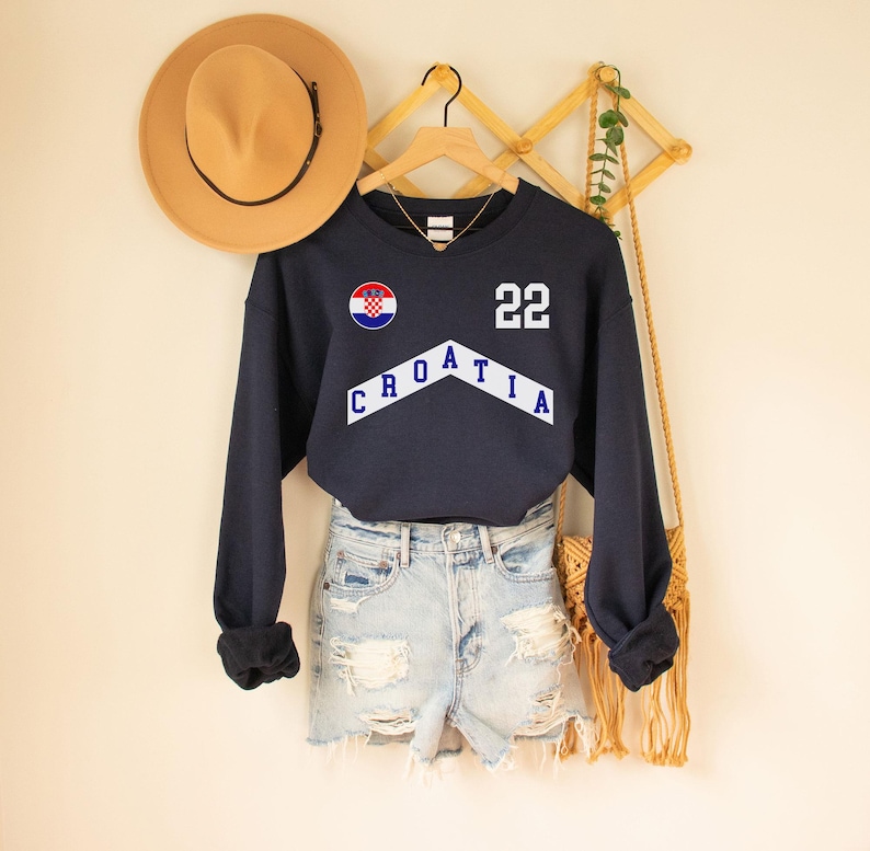 May include: A navy blue sweatshirt with a white chevron design that says "CROATIA" and a white number "22". The sweatshirt also features a circular patch with the Croatian flag.