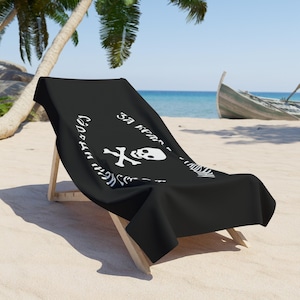 May include: Black beach towel draped over a wooden beach chair. The towel features a white skull and crossbones design with text. The chair is on a sandy beach with a boat in the background.