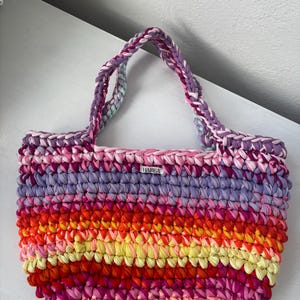 May include: A handmade, colorful tote bag with a striped pattern in shades of pink, purple, orange, and yellow. The bag has braided handles and a small tag that reads "HAMBA".