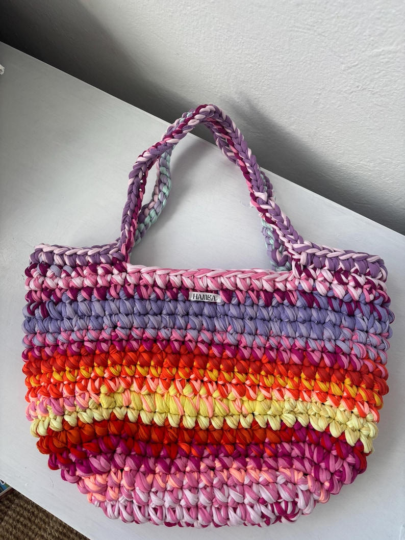May include: A handmade, colorful tote bag with a striped pattern in shades of pink, purple, orange, and yellow. The bag has braided handles and a small tag that reads "HAMBA".