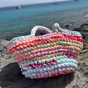 May include: A colorful, handmade tote bag with a woven design. The bag features a variety of colors, including pink, orange, yellow, and green. The bag has two handles and a label that reads "HANBA". The bag is sitting on a rock near the ocean.