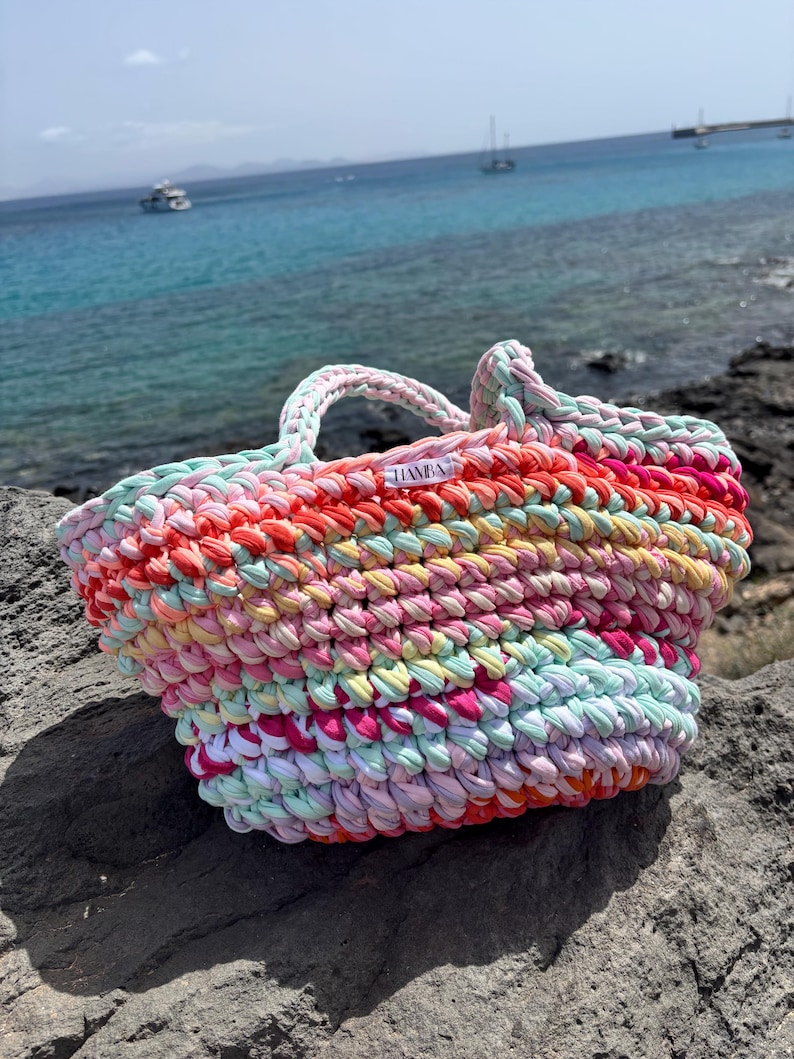 May include: A colorful, handmade tote bag with a woven design. The bag features a variety of colors, including pink, orange, yellow, and green. The bag has two handles and a label that reads "HANBA". The bag is sitting on a rock near the ocean.