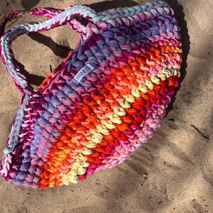 May include: A colorful, handmade tote bag rests on sand. The bag is crafted from thick, braided fabric in a vibrant array of colors including pink, orange, yellow, and purple. The handles are braided and the bag has a label that says "HANBA".