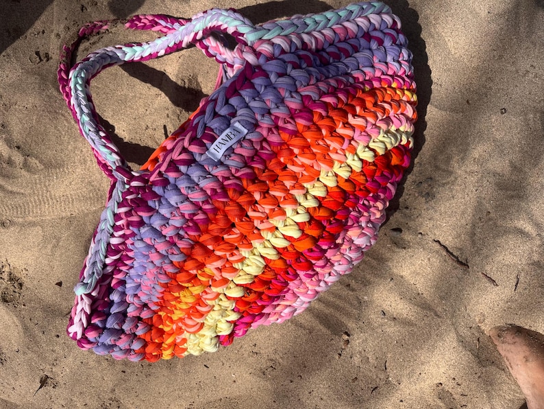 May include: A colorful, handmade tote bag rests on sand. The bag is crafted from thick, braided fabric in a vibrant array of colors including pink, orange, yellow, and purple. The handles are braided and the bag has a label that says "HANBA".