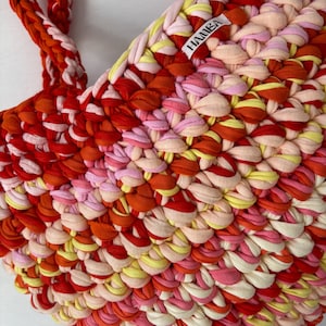 May include: A handmade tote bag in a vibrant mix of red, orange, pink, and yellow fabric strips. The bag features braided handles and a textured, woven design. The label "HAMBA" is visible on the side of the bag.