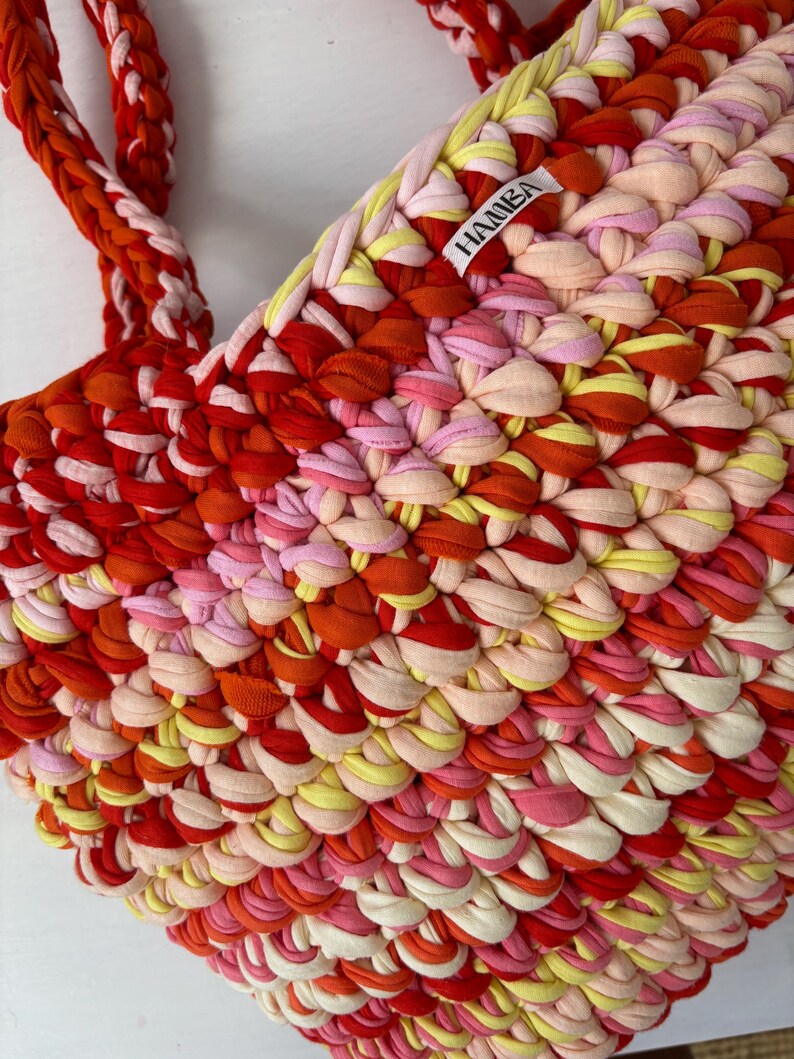 May include: A handmade tote bag in a vibrant mix of red, orange, pink, and yellow fabric strips. The bag features braided handles and a textured, woven design. The label "HAMBA" is visible on the side of the bag.