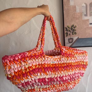 May include: A vibrant, hand-crocheted tote bag in shades of pink, orange, and cream. The bag features sturdy, braided handles and a rectangular shape. A small white tag with the word "HAME" is visible. This colorful bag is perfect for carrying essentials.
