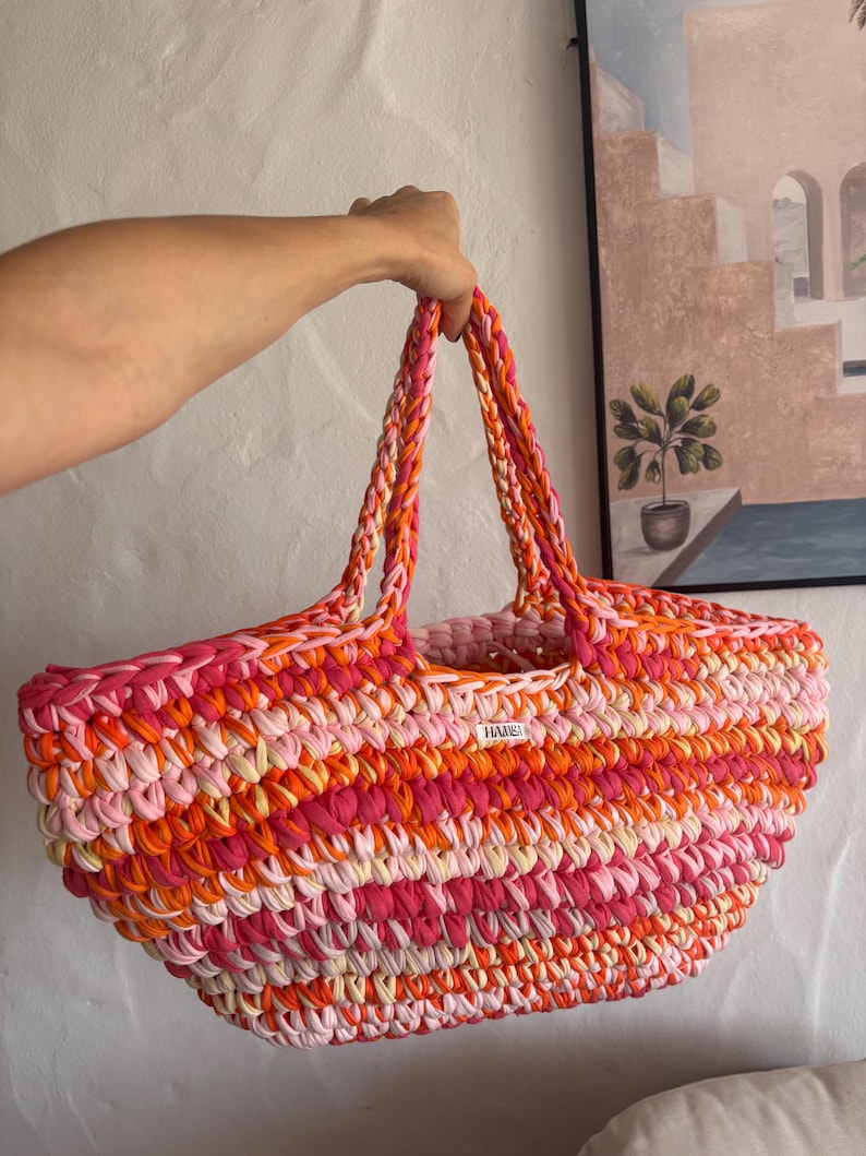 May include: A vibrant, hand-crocheted tote bag in shades of pink, orange, and cream. The bag features sturdy, braided handles and a rectangular shape. A small white tag with the word "HAME" is visible. This colorful bag is perfect for carrying essentials.