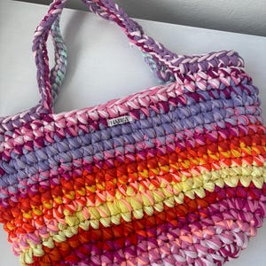 May include: A handmade, colorful tote bag with braided handles. The bag features a vibrant mix of pink, purple, orange, and yellow fabric strips, creating a striped pattern. The label on the bag reads "HAMBA".