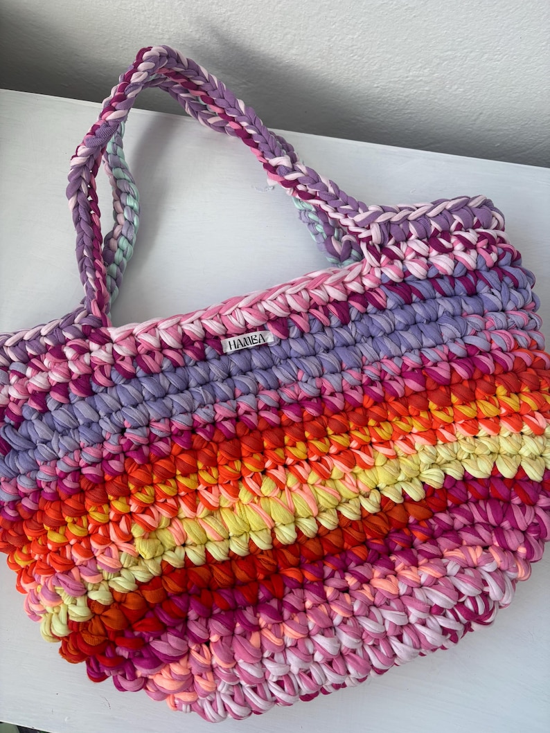 May include: A handmade, colorful tote bag with braided handles. The bag features a vibrant mix of pink, purple, orange, and yellow fabric strips, creating a striped pattern. The label on the bag reads "HAMBA".