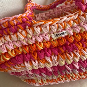 May include: A handmade, colorful tote bag with a braided handle. The bag is crafted from thick, knitted fabric in shades of pink, orange, and cream. The label on the bag reads "HAMBA".