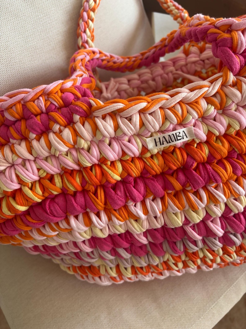 May include: A handmade, colorful tote bag with a braided handle. The bag is crafted from thick, knitted fabric in shades of pink, orange, and cream. The label on the bag reads "HAMBA".