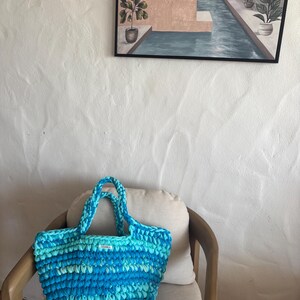 May include: A handmade, blue and turquoise crochet tote bag with two handles. The bag is sitting on a chair with a cream-colored cushion. A painting of a pool scene hangs on the white wall behind the chair.