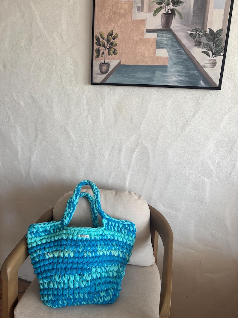 May include: A handmade, blue and turquoise crochet tote bag with two handles. The bag is sitting on a chair with a cream-colored cushion. A painting of a pool scene hangs on the white wall behind the chair.