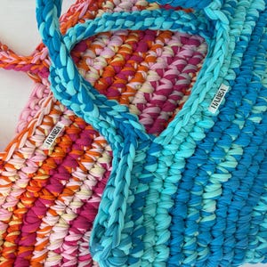 May include: Two handmade crochet tote bags in vibrant colors. One bag features a mix of pink, orange, and yellow, while the other is a gradient of blues. Both have braided handles and a small label that reads "HAMISA".