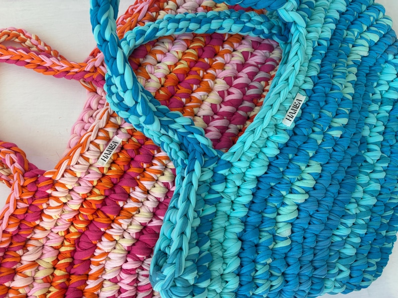May include: Two handmade crochet tote bags in vibrant colors. One bag features a mix of pink, orange, and yellow, while the other is a gradient of blues. Both have braided handles and a small label that reads "HAMISA".
