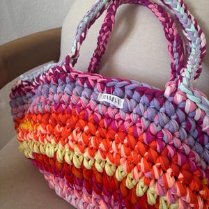 May include: A handmade tote bag with a vibrant, multicolored design. The bag features a woven pattern with shades of pink, orange, yellow, and purple. The handles are braided in coordinating colors. A small white label with the text "HAMBA" is visible.