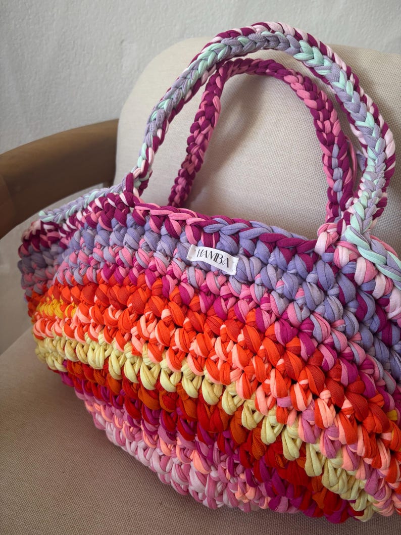 May include: A handmade tote bag with a vibrant, multicolored design. The bag features a woven pattern with shades of pink, orange, yellow, and purple. The handles are braided in coordinating colors. A small white label with the text "HAMBA" is visible.