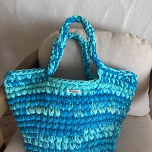 May include: A handmade, blue and turquoise crochet tote bag with sturdy handles. The bag features a textured stitch pattern and a small white label with the text "HAMEA". The bag is sitting on a neutral-colored cushion.