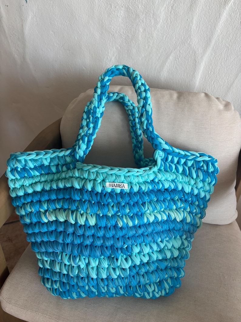 May include: A handmade, blue and turquoise crochet tote bag with sturdy handles. The bag features a textured stitch pattern and a small white label with the text "HAMEA". The bag is sitting on a neutral-colored cushion.