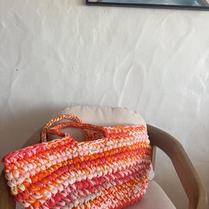 May include: A large, rectangular crochet tote bag in shades of pink, orange, and white. The bag has braided handles and a label on the front. The bag is sitting on a beige cushion on a wooden chair.