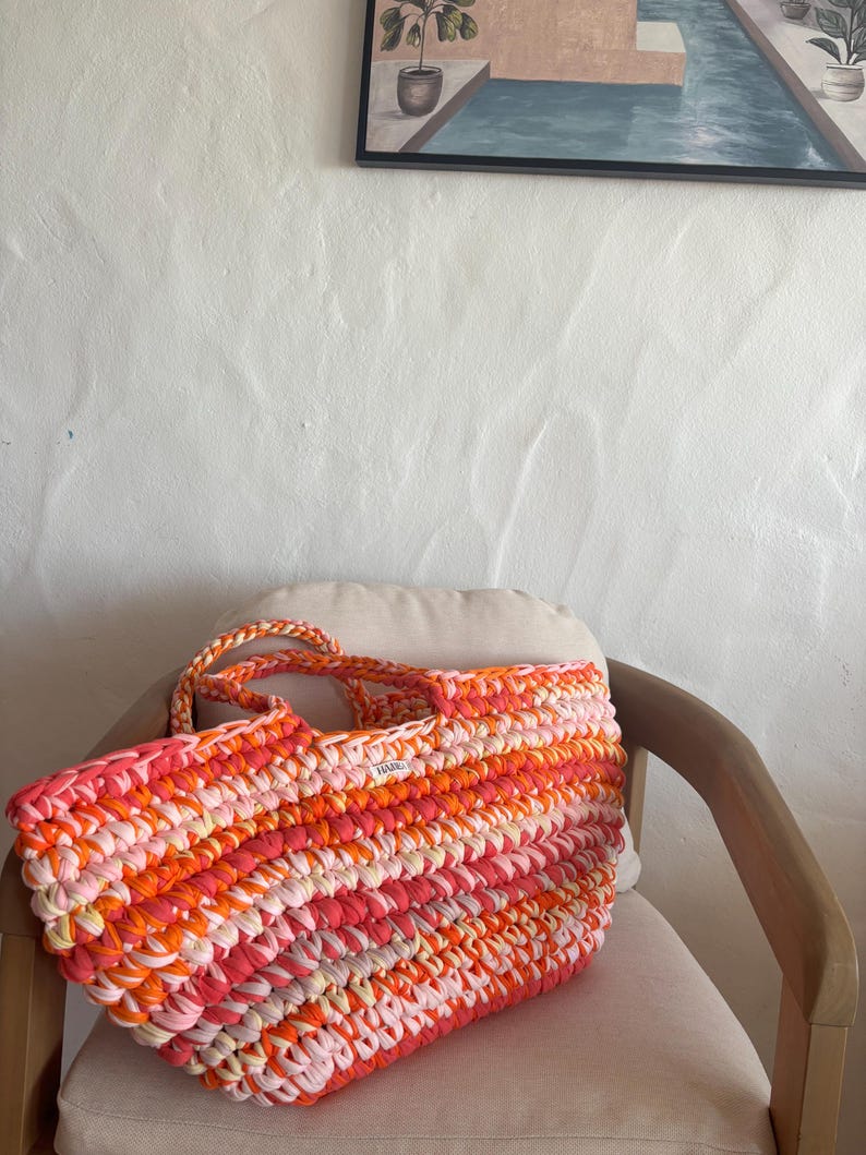 May include: A large, rectangular crochet tote bag in shades of pink, orange, and white. The bag has braided handles and a label on the front. The bag is sitting on a beige cushion on a wooden chair.