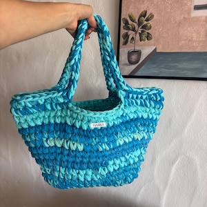 May include: A handmade, blue and turquoise crochet tote bag with braided handles. The bag features a label that reads "HAMSA". The bag is made of a soft, textured material and is designed for carrying items.