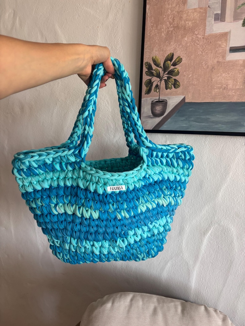 May include: A handmade, blue and turquoise crochet tote bag with braided handles. The bag features a label that reads "HAMSA". The bag is made of a soft, textured material and is designed for carrying items.