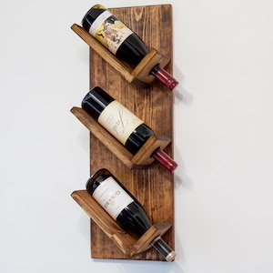 May include: A wooden wine rack with three angled slots, each holding a wine bottle. The rack is made of dark stained wood and is mounted on a white wall. The wine bottles have red and white labels.