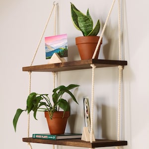 May include: Two wooden shelves suspended from the ceiling with rope. The top shelf features a potted plant and a photograph of mountains. The bottom shelf displays a potted plant, a book titled "House Plants", and a photograph.