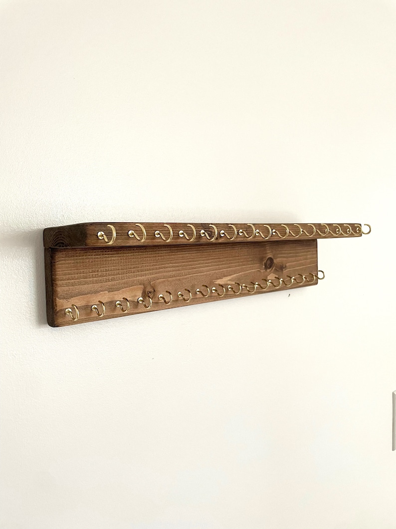 May include: A wooden key holder with a shelf and multiple gold hooks. The key holder is mounted on a white wall.